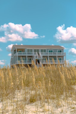 A large, white beach house elevated on stilts sits behind a patch of tall, golden beach grass. The sky above is bright blue with a few fluffy white clouds scattered across.