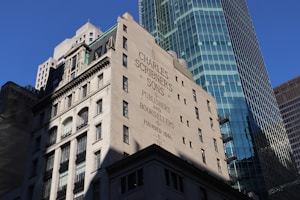 A historic building with the name 'Charles Scribner's Sons' and the words 'Publishers and Booksellers Founded 1846' painted on its wall. The architecture features classic elements with multiple windows and decorative stonework. In the background, a modern skyscraper with a glass facade rises, reflecting the blue sky.