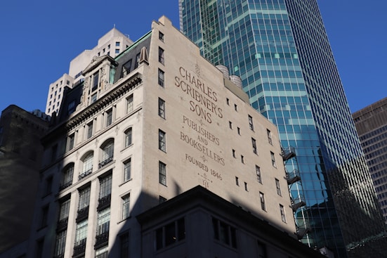 A historic building with the name 'Charles Scribner's Sons' and the words 'Publishers and Booksellers Founded 1846' painted on its wall. The architecture features classic elements with multiple windows and decorative stonework. In the background, a modern skyscraper with a glass facade rises, reflecting the blue sky.