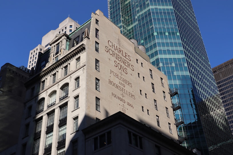 A historic building with the name 'Charles Scribner's Sons' and the words 'Publishers and Booksellers Founded 1846' painted on its wall. The architecture features classic elements with multiple windows and decorative stonework. In the background, a modern skyscraper with a glass facade rises, reflecting the blue sky.