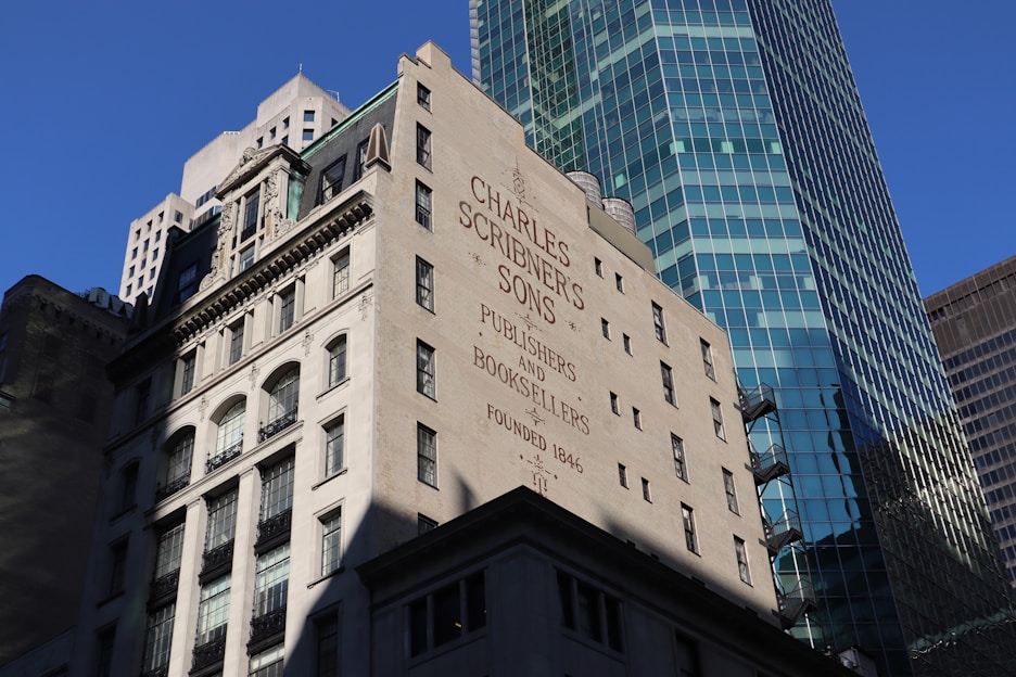 A historic building with the name 'Charles Scribner's Sons' and the words 'Publishers and Booksellers Founded 1846' painted on its wall. The architecture features classic elements with multiple windows and decorative stonework. In the background, a modern skyscraper with a glass facade rises, reflecting the blue sky.