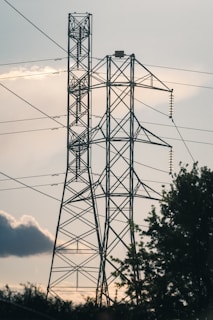 Two large metal electricity pylons stand tall against the backdrop of a cloudy sky. The pylons are interconnected with multiple power lines, and there is some foliage visible at the base, indicating a natural setting. The light suggests early evening or late afternoon, casting shadows and creating a calm atmosphere.
