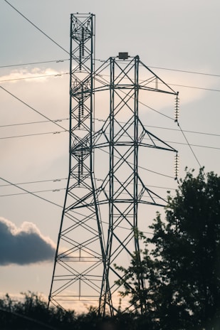 Two large metal electricity pylons stand tall against the backdrop of a cloudy sky. The pylons are interconnected with multiple power lines, and there is some foliage visible at the base, indicating a natural setting. The light suggests early evening or late afternoon, casting shadows and creating a calm atmosphere.