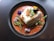 A beautifully plated dish featuring a rectangular piece of white fish garnished with fried garlic slices, microgreens, and edible flowers. The fish is surrounded by a bright orange puree or sauce, adding a contrasting color to the composition.