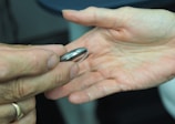 Two hands are captured in close-up. One hand, on the left, holds a metallic ring-like object between the thumb and index finger. The other hand, open with the palm facing up, is positioned as if ready to receive the object.