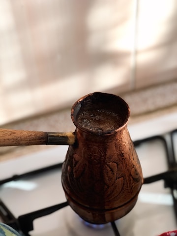 Elegant copper coffee pot steaming beside a cream-colored ceramic cup on a dark brown wooden table.