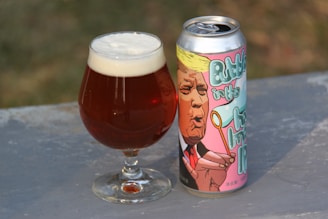 A glass of beer with a thick head of foam sits next to a colorful, illustrated beer can. The can features an illustration of a person blowing a party horn, rendered in a cartoon style with pastel colors.