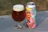 A glass of beer with a thick head of foam sits next to a colorful, illustrated beer can. The can features an illustration of a person blowing a party horn, rendered in a cartoon style with pastel colors.
