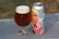 A glass of beer with a thick head of foam sits next to a colorful, illustrated beer can. The can features an illustration of a person blowing a party horn, rendered in a cartoon style with pastel colors.