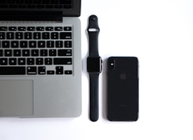 A silver laptop with a black keyboard is open and positioned on a white surface. To its right, a black smartwatch with a strap is placed beside a black smartphone. The smartwatch and smartphone both display a minimalistic design with no visible screens active.