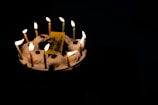 A cake with fourteen lit candles arranged around the perimeter. The cake appears to have white frosting with red cherry-like decorations on top and a yellow rectangular piece with text. The background is dark, enhancing the glow of the candles.