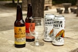 Several bottles and cans of beverages are placed on a wooden surface outdoors. The arrangement includes two glass bottles labeled as ginger beer and two metallic cans branded as China White Belgian Witbier. An empty glass is positioned in front of the beverages. The background shows a blurred outdoor setting.