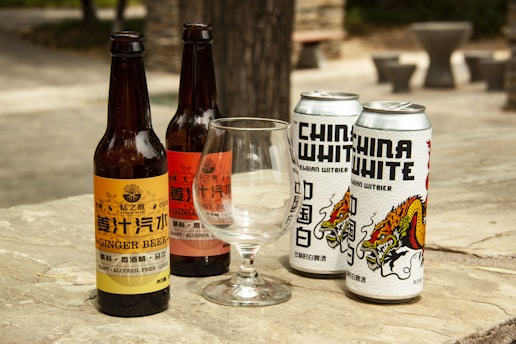 Several bottles and cans of beverages are placed on a wooden surface outdoors. The arrangement includes two glass bottles labeled as ginger beer and two metallic cans branded as China White Belgian Witbier. An empty glass is positioned in front of the beverages. The background shows a blurred outdoor setting.