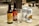 Several bottles and cans of beverages are placed on a wooden surface outdoors. The arrangement includes two glass bottles labeled as ginger beer and two metallic cans branded as China White Belgian Witbier. An empty glass is positioned in front of the beverages. The background shows a blurred outdoor setting.