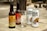 Several bottles and cans of beverages are placed on a wooden surface outdoors. The arrangement includes two glass bottles labeled as ginger beer and two metallic cans branded as China White Belgian Witbier. An empty glass is positioned in front of the beverages. The background shows a blurred outdoor setting.