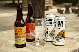 Several bottles and cans of beverages are placed on a wooden surface outdoors. The arrangement includes two glass bottles labeled as ginger beer and two metallic cans branded as China White Belgian Witbier. An empty glass is positioned in front of the beverages. The background shows a blurred outdoor setting.