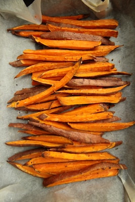 Numerous baked sweet potato fries are arranged on a baking sheet lined with parchment paper. The fries are cut into thick, rustic slices, showcasing a vibrant orange interior and slightly browned edges.