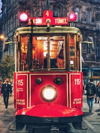 A vintage red tram is prominently featured, running through a bustling urban area with pedestrians walking along the street. The tram displays signs for 'Taksim' and 'Tünel' and gives off a warm glow from its lights. The surrounding buildings have an old architectural style that adds to the nostalgic atmosphere.