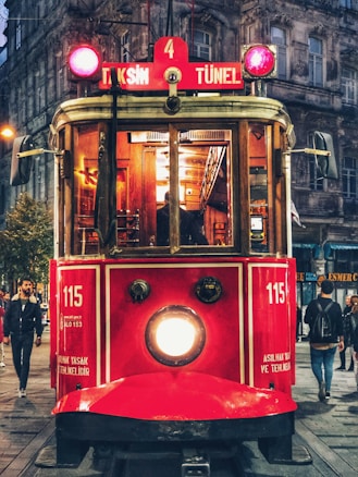 A vintage red tram is prominently featured, running through a bustling urban area with pedestrians walking along the street. The tram displays signs for 'Taksim' and 'Tünel' and gives off a warm glow from its lights. The surrounding buildings have an old architectural style that adds to the nostalgic atmosphere.