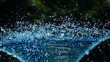 A dynamic scene of water droplets captured mid-air with a dark, blurred background. The droplets create a sense of movement and sparkle in the light, highlighting their clarity and fluidity.