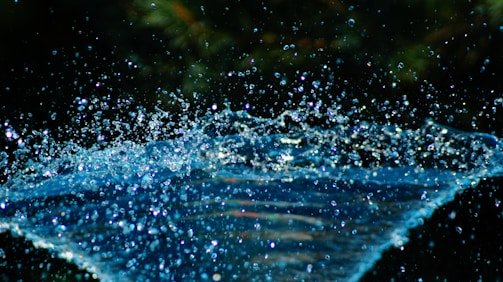 A dynamic scene of water droplets captured mid-air with a dark, blurred background. The droplets create a sense of movement and sparkle in the light, highlighting their clarity and fluidity.