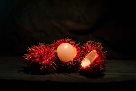 Three rambutan fruits are displayed, with one fruit peeled open to reveal the white, translucent flesh inside. The fruits have a bright red, hairy exterior and are set against a dark background, enhancing the vibrant color of the rambutan.