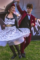 A man and woman are dancing energetically on a grassy area. The woman has red hair and is wearing a patterned dress and a black leather jacket, while the man is dressed in a red suit with a bow tie. The background features a banner with large letters, possibly part of an outdoor event or festival setup.