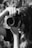 A black and white photograph capturing a person holding a large camera close to their face, with long hair cascading over their shoulders. The person's hand is prominently grasping the lens, and the background is softly blurred, adding focus to the camera and photographer.