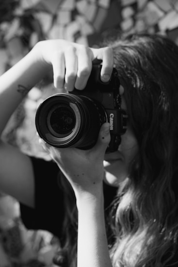A black and white photograph capturing a person holding a large camera close to their face, with long hair cascading over their shoulders. The person's hand is prominently grasping the lens, and the background is softly blurred, adding focus to the camera and photographer.