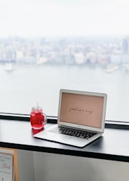 A laptop displaying the phrase 'good vibes only' is placed on a black windowsill. Next to it is a red beverage in a mason jar with a straw. The background features a blurred cityscape view through a large window, suggesting a high-rise perspective.