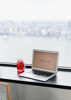 A laptop displaying the phrase 'good vibes only' is placed on a black windowsill. Next to it is a red beverage in a mason jar with a straw. The background features a blurred cityscape view through a large window, suggesting a high-rise perspective.