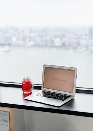 A laptop displaying the phrase 'good vibes only' is placed on a black windowsill. Next to it is a red beverage in a mason jar with a straw. The background features a blurred cityscape view through a large window, suggesting a high-rise perspective.