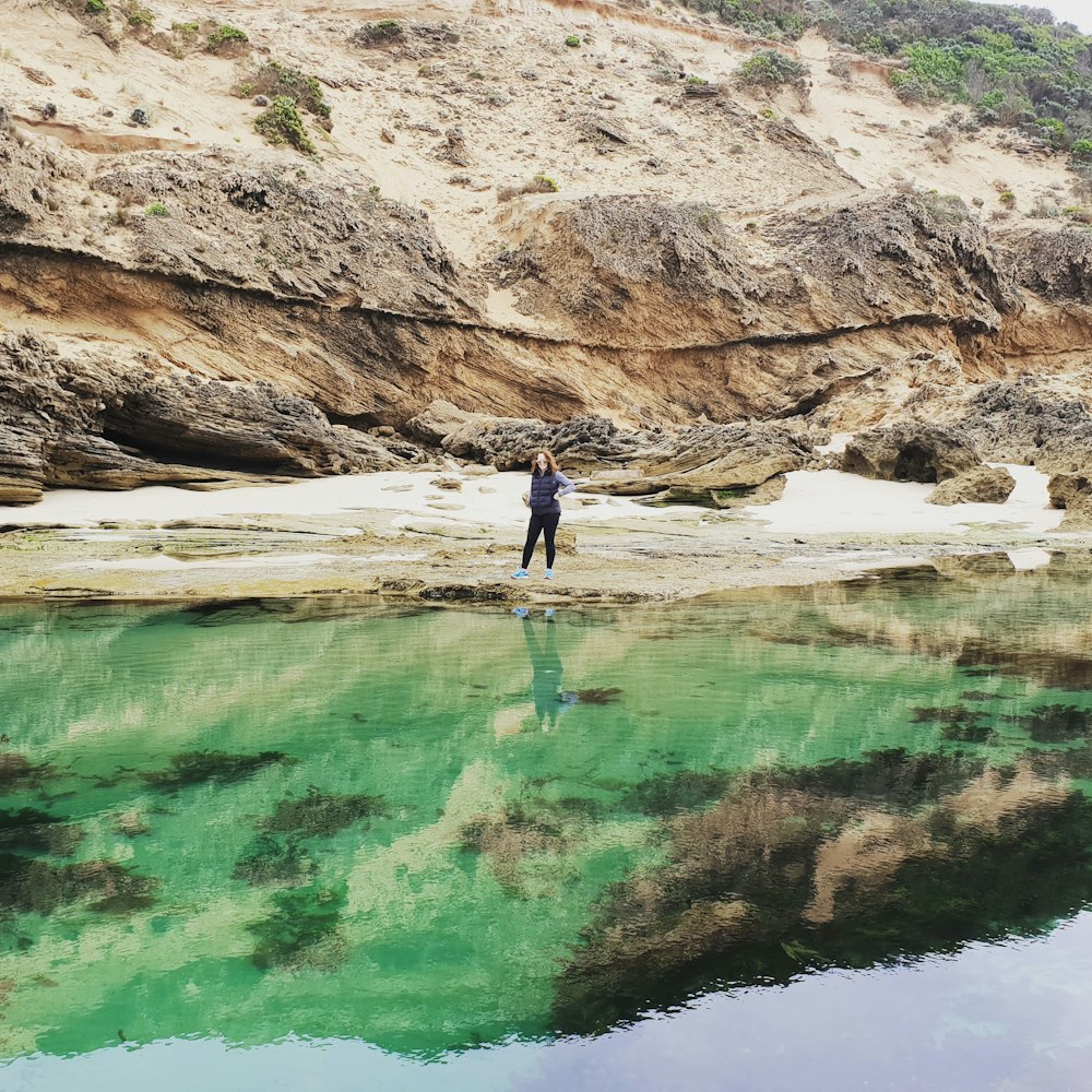 person in black shirt and black pants standing on brown rock formation near body of water