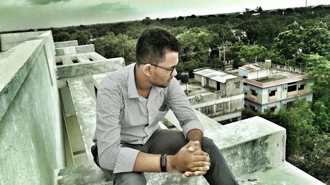 A man sits pensively on the edge of a rooftop, overlooking a suburban area with trees and scattered buildings. He is wearing a striped shirt and glasses, resting his arms on his knees. The sky is overcast, contributing to a subdued atmosphere.