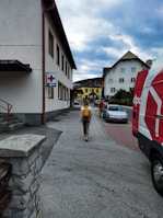 A person carrying a yellow backpack is walking along a sidewalk in a residential area. Several buildings are visible, including one with a sign featuring a red cross and an arrow. A red and white van is parked nearby, and a couple of cars can be seen on the street. The sky is overcast.