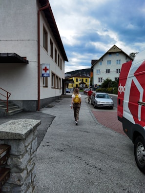 A person carrying a yellow backpack is walking along a sidewalk in a residential area. Several buildings are visible, including one with a sign featuring a red cross and an arrow. A red and white van is parked nearby, and a couple of cars can be seen on the street. The sky is overcast.