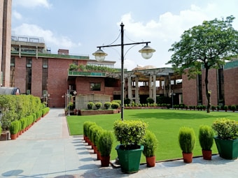 A well-maintained garden with neatly trimmed grass and potted plants lines a paved walkway, leading to a large brick building. The architecture is complemented by tall trees and vintage-style lampposts. The atmosphere appears serene and orderly, with the garden adding a touch of nature to the urban setting.