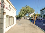 A sunny urban street scene with a wide sidewalk and a building on the left displaying a sign for a property management company. Trees with lush green foliage provide shade further down the street. Several vehicles, including cars and a truck, are parked or driving on the road. A sign indicating a loading zone is visible, and the sky is clear and blue.
