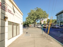 A sunny urban street scene with a wide sidewalk and a building on the left displaying a sign for a property management company. Trees with lush green foliage provide shade further down the street. Several vehicles, including cars and a truck, are parked or driving on the road. A sign indicating a loading zone is visible, and the sky is clear and blue.