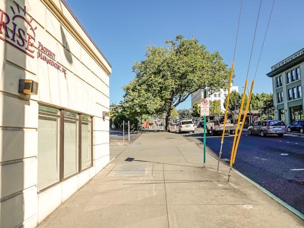 A sunny urban street scene with a wide sidewalk and a building on the left displaying a sign for a property management company. Trees with lush green foliage provide shade further down the street. Several vehicles, including cars and a truck, are parked or driving on the road. A sign indicating a loading zone is visible, and the sky is clear and blue.