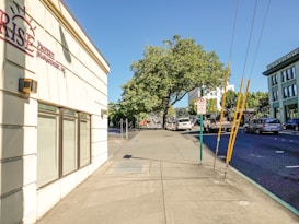 A sunny urban street scene with a wide sidewalk and a building on the left displaying a sign for a property management company. Trees with lush green foliage provide shade further down the street. Several vehicles, including cars and a truck, are parked or driving on the road. A sign indicating a loading zone is visible, and the sky is clear and blue.