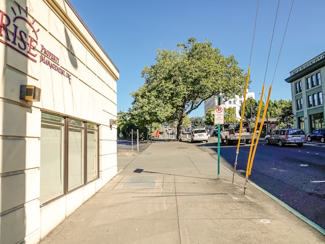 A sunny urban street scene with a wide sidewalk and a building on the left displaying a sign for a property management company. Trees with lush green foliage provide shade further down the street. Several vehicles, including cars and a truck, are parked or driving on the road. A sign indicating a loading zone is visible, and the sky is clear and blue.