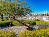 A large metal anchor is displayed on a bed of pebbles surrounded by neatly trimmed bushes. The setting is a waterfront park with a marina full of sailboats and yachts in the background. Trees provide shade over a path with benches, while a lamppost stands nearby. A wooden sign dedicated to fishermen is visible beside the anchor.