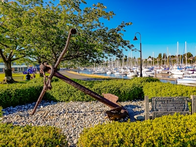 A large metal anchor is displayed on a bed of pebbles surrounded by neatly trimmed bushes. The setting is a waterfront park with a marina full of sailboats and yachts in the background. Trees provide shade over a path with benches, while a lamppost stands nearby. A wooden sign dedicated to fishermen is visible beside the anchor.