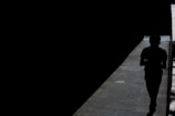 A silhouette of a person walking through a narrow pathway illuminated by sunlight, creating a stark contrast between the brightly lit path and the surrounding darkness.