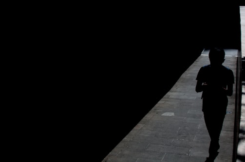 A silhouette of a person walking through a narrow pathway illuminated by sunlight, creating a stark contrast between the brightly lit path and the surrounding darkness.