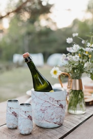 A wine bottle is chilling in a marble-patterned cooler on a wooden table. Two matching tumblers are nearby. A vase with wildflowers, including daisies, is in the background, alongside a copper pitcher. The setting appears to be outdoors with greenery in the backdrop, suggesting a picnic or outdoor gathering.