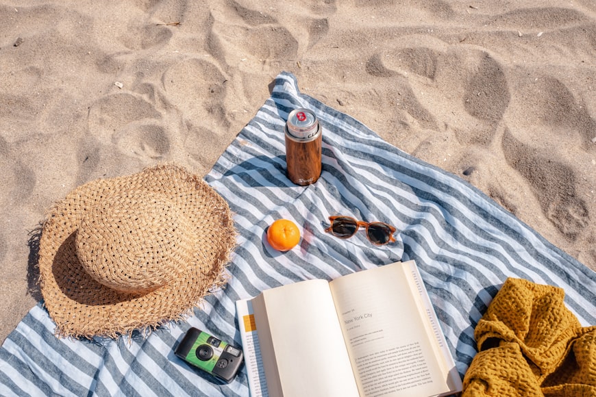 A beach with a blanket spread out with a hat, sunglasses, an orange, a drink and a book on it. 