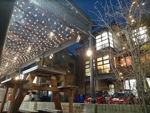 A cozy evening scene under the large wooden gazebo with picnic benches and string lights glowing warmly.