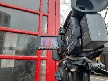A digital video camera on a tripod is positioned in front of a red painted wooden structure with glass panes, likely a door or window. The camera's LCD screen displays the red structure, indicating that the camera is recording or preparing to record the scene. There are some reflections in the glass, and partial visibility of trees in the background.
